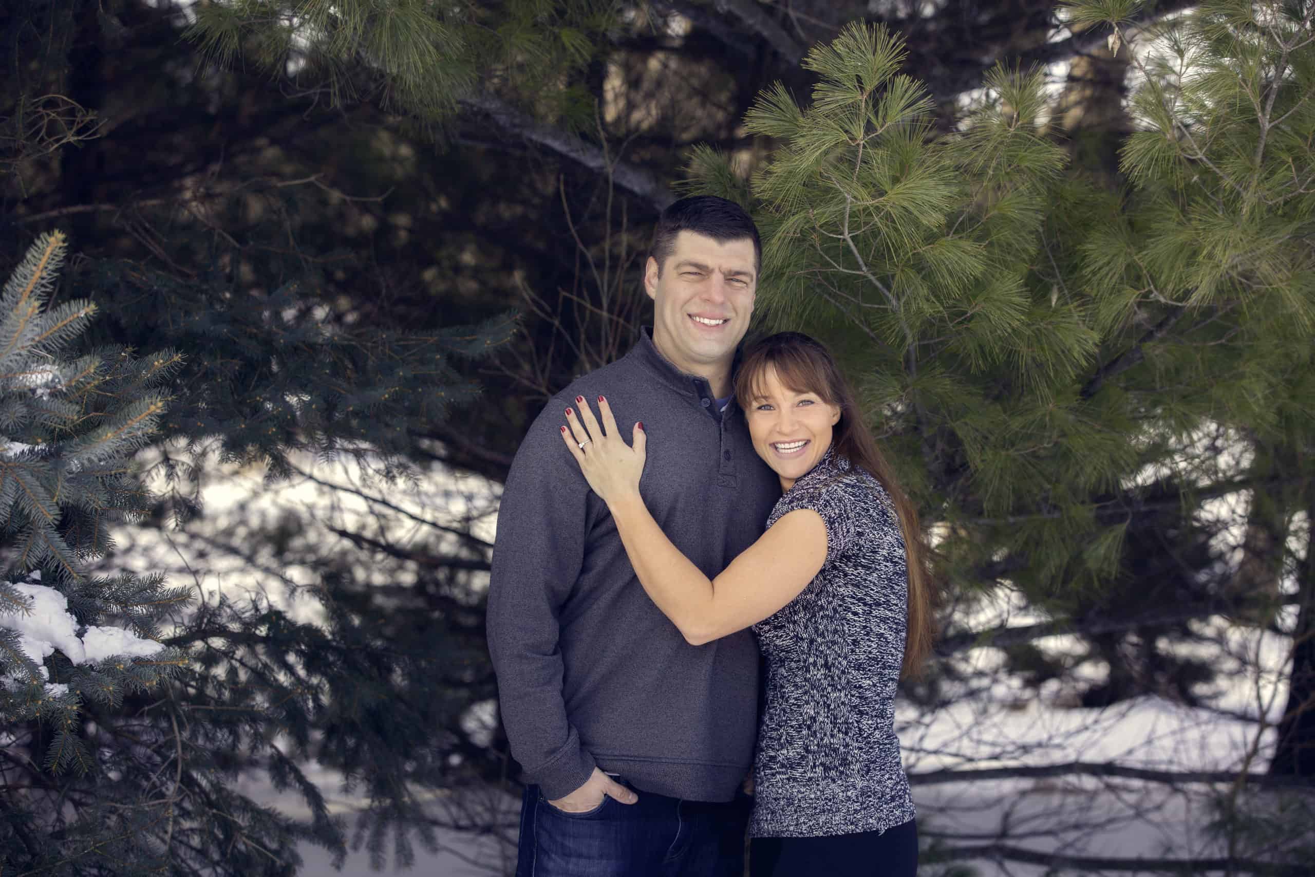 Couple stands in front of trees and woman puts her left hand on man's chest.