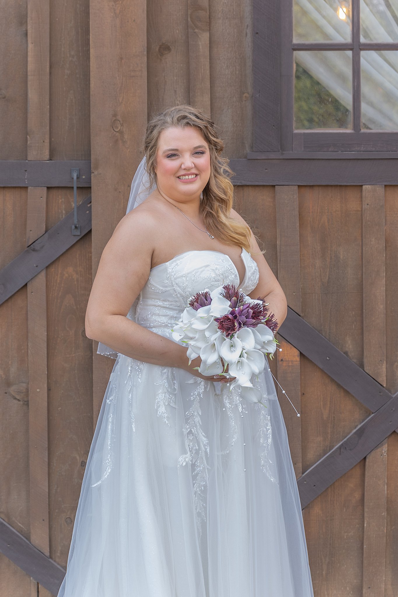 bride stands in front of barn holding her white and purple bouquet