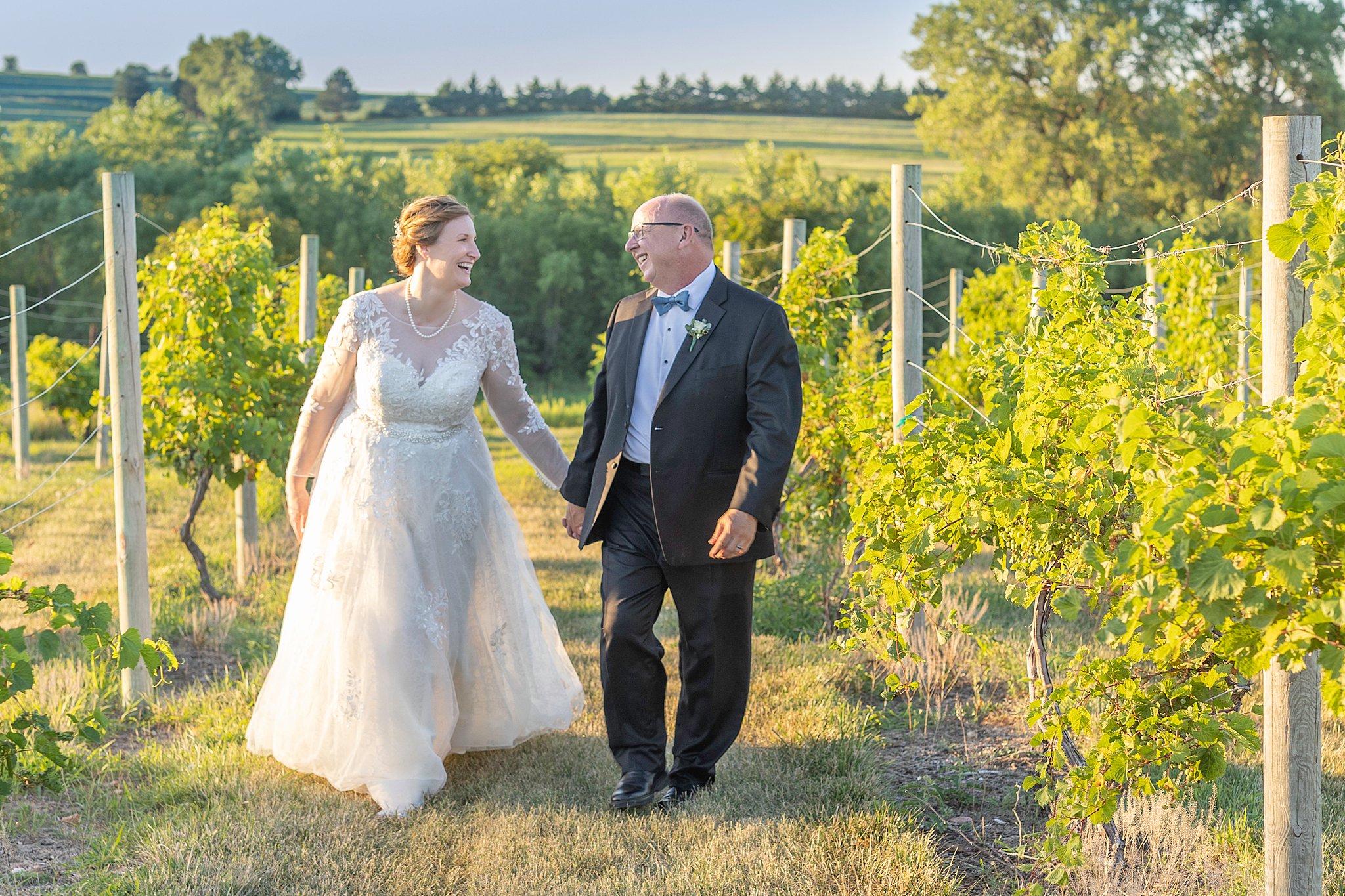 Newlyweds walk down a row of a vineyard holding hands with rolling hills behind them