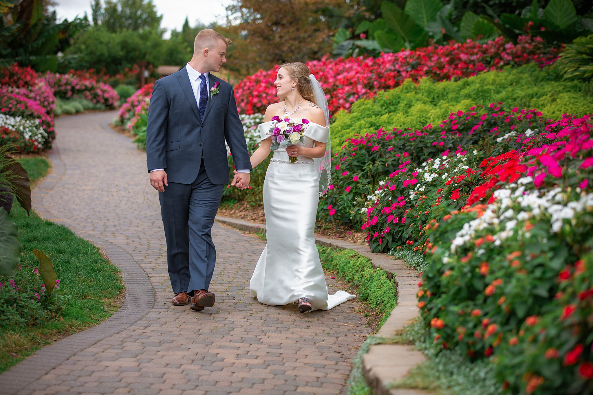 A newlywed couple holds hands while walking through a colorful flower garden path in a blue suit and silk wedding dress sunken gardens wedding lincoln ne