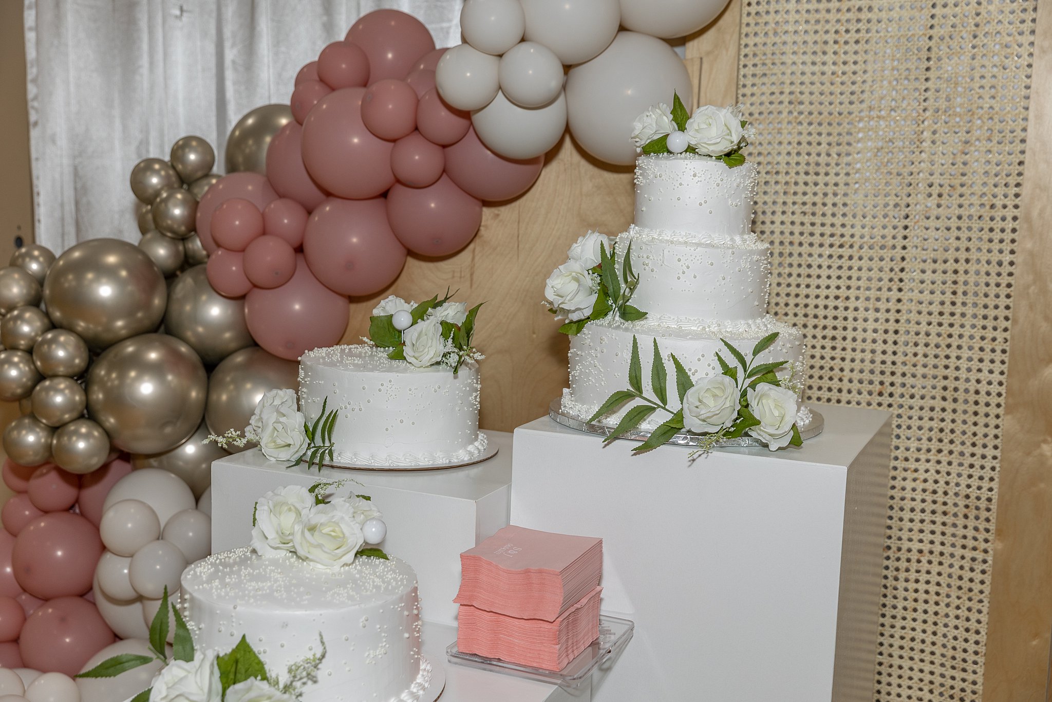 A three-tier white wedding cake sits on a white cube with white roses as decorations next to two other similar cakes the gala lincoln ne