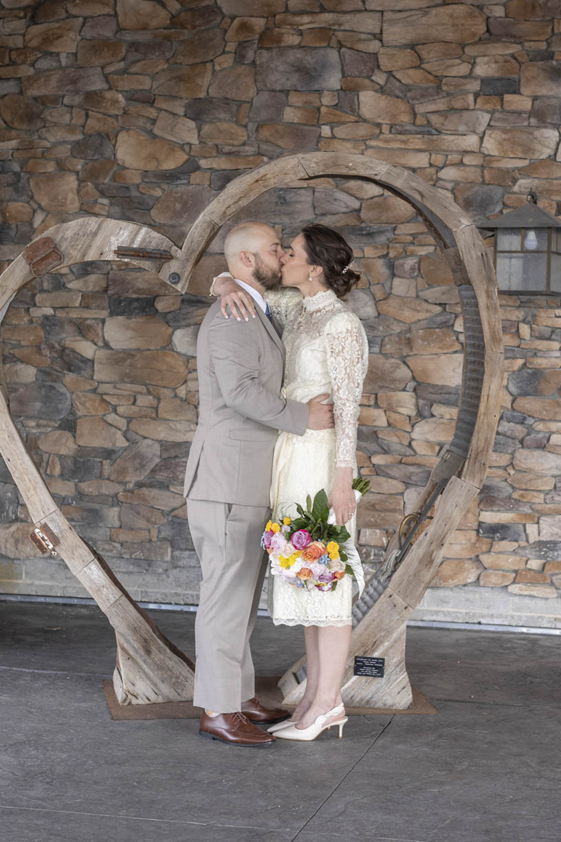 newlyweds kiss in front of a rustic wooden heart and stone wall Wilderness Ridge wedding