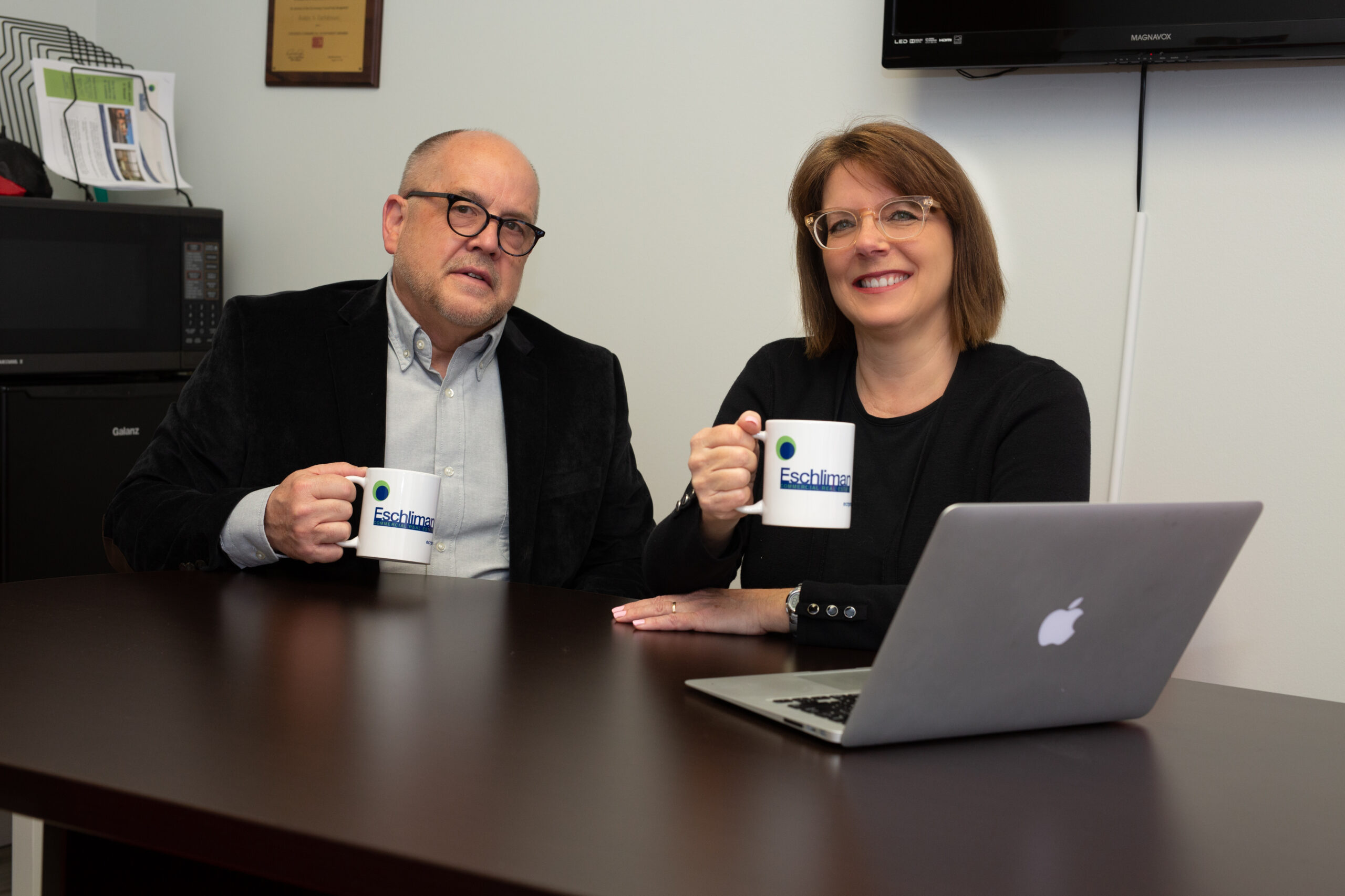 Couple sit at desk holding mugs with laptop on table